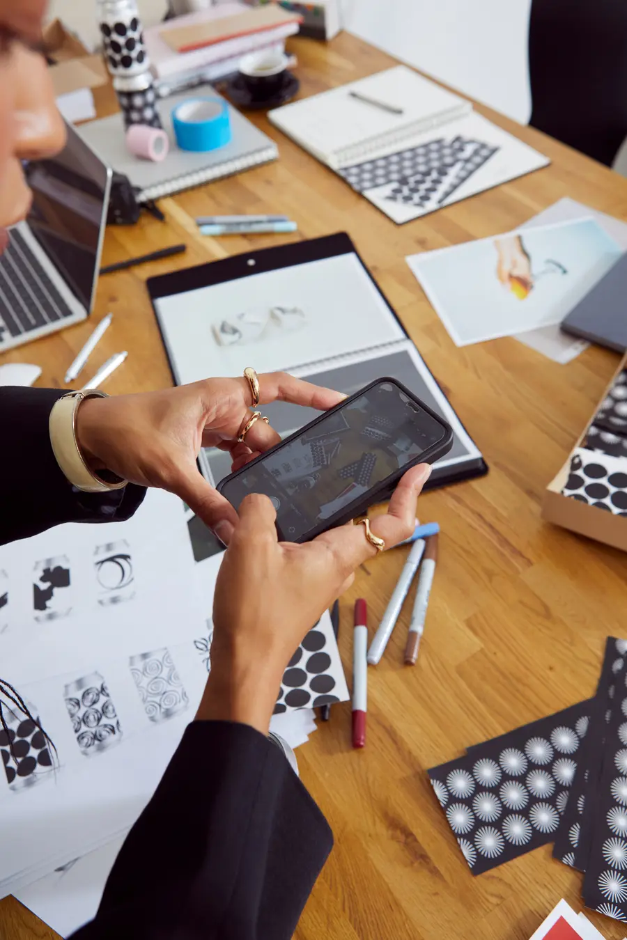 Hands using a smartphone above a desk with a laptop, printouts, and markers